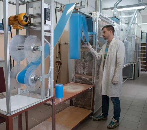 A worker in a lab coat at a tall frame unit feeding blue abrasive belt over rollers